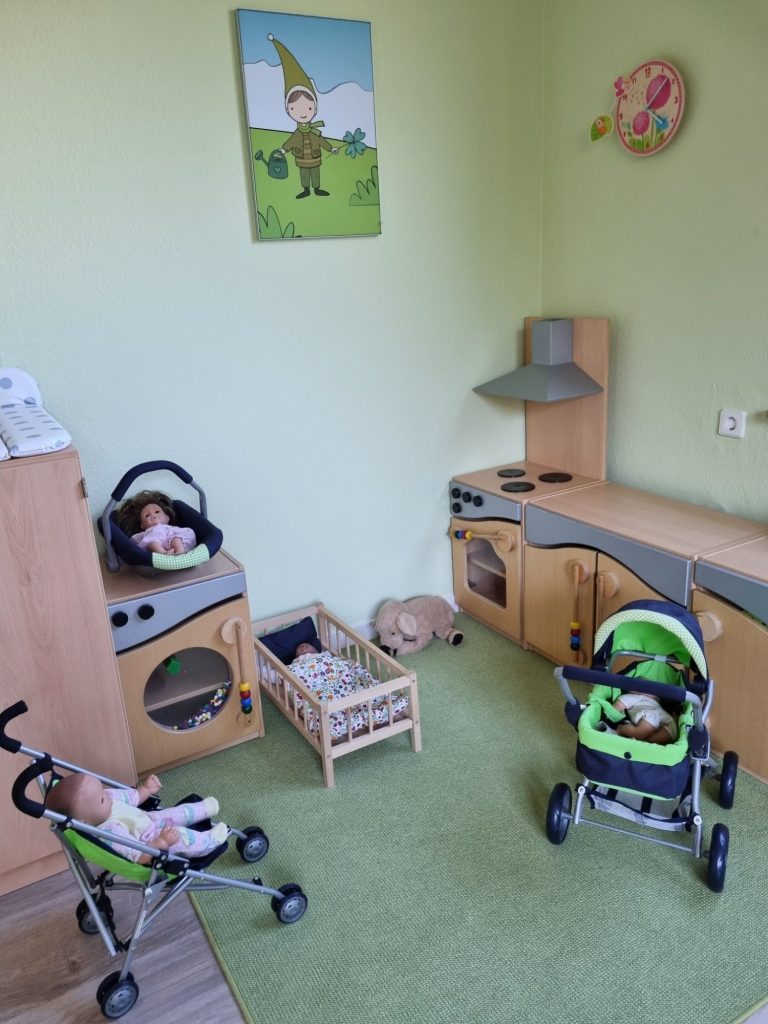 Eine Puppenecke mit Kinderküche aus Holz, Puppenwagen, einem Puppenbett und mehreren Puppen auf grünem Teppich.